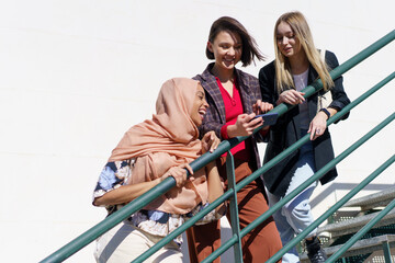 Diverse cheerful female students smiling while sharing smartphone on staircase