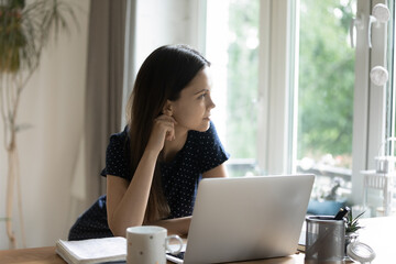Young thoughtful attractive woman sit at table with laptop looks aside, finished studying on computer resting staring out window, ponders over ideas or task, distracted from work, daydreaming at home
