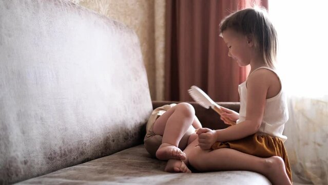 Funny children siblings play together on the sofa. Toddler and baby in a reusable diaper at home in a real interior. Sibling relationships. Sister combing and hugging little brother