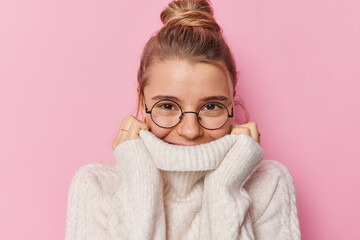 Beautiful young woman with fair hair gathered in bun hides mouth behind collar of sweater wears...