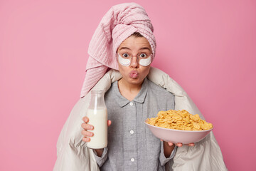 Surprised young woman has healthy breakfast holds bowl of cereals and milk bottle wears pajama and beauty patches bath towel covered with blanket keeps lips folded isolated over pink background