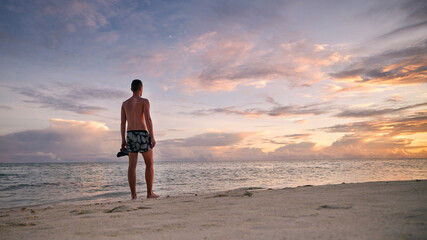 Rear view of young man on idyllic white sand beach. Tourist in swimwear watching colorful sunset in...