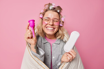 Cheerful woman applies hair rollers for making hairstyle wears pajama wrapped in blanket chooses between menstruation cup and gasket during periods smiles gladfully isolated over pink background