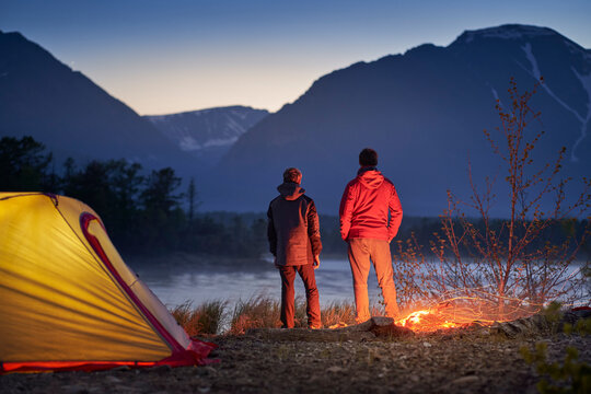 Two tourists standing at tent with campfire and looking at the mountain. Camping in wilderness area