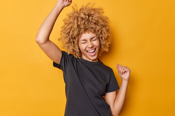 Emotional curly haired woman clenches fists in air feels excited exclaims from joy wears casual black t shirt isolated over yellow background celebrates succes or achievement. Yes I did it finally