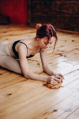 Attractive athletic young woman with red hair doing yoga, stretching and training, in a sporty black bra and beige leggings, in the gym, studio with a ballet machine..