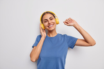 Waist up shot of positive carefree European woman dances and catches every bit of music wears casual blue t shirt listens favorite song via wireless headphones isolated over white background.