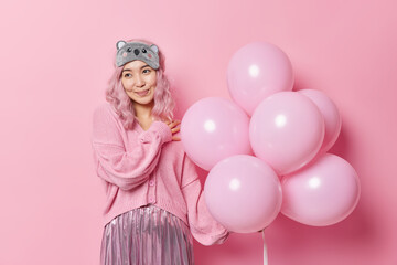 Obraz na płótnie Canvas Dreamy lovely young Asian woman with long dyed hair dressed in festive clothes holds bunch of inflated balloons enjoys party time and holiday isolated over pink background. Holidays concept.