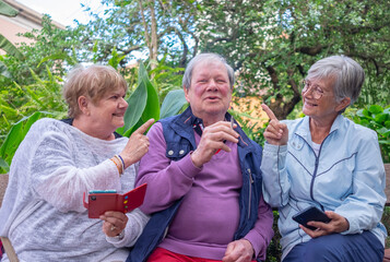 Happy group of three seniors sitting relaxed on the park bench. The man smokes a cigarette, the women hold the cell phone asking the man not to smoke