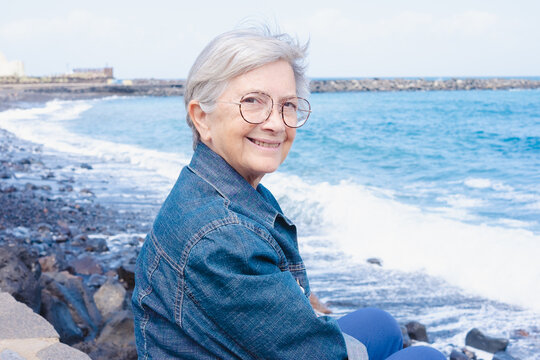 Portrait of white-haired smiling senior woman sitting on the beach wearing eyeglasses and denim jacket. Attractive 70 years old woman enjoying good time and vacation