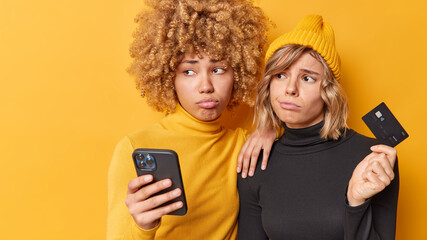 Upset young two women have sad expressions use mobile phone and credit card cannot use application dressed casually isolated over yellow background look away. Hard money transfer online payment