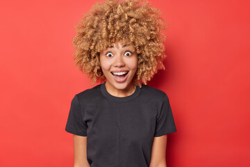 Blonde curly haired woman looks with happy surprised expression smiles broadly dressed in casual black t shirt isolated over bright red background reacts on unexpected offer. Human reactions