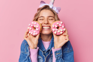 Cheerful fair haired young European woman keeps eyes closed smiles toothily holds two donuts with marshmallow glad to eat favorite dessert dressed in denim jacket isolated over pink background