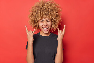 Positive young curly woman shows horn gesture enjoys rock music wears black casual t shirt isolated...