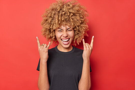 Positive young curly woman shows horn gesture enjoys rock music wears black casual t shirt isolated over vivid red background. Overjoyed curly haired female model demonstrates heavy metal sign