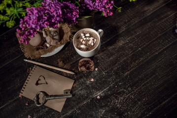 Hot chocolate (cocoa) in a glass mug with two tubes for drinking.There are some macarons on a linen napkin on the wood table with some lilac
