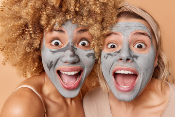 Horizontal shot of amazed surprised women apply facial clay masks undergo beauty procedures react on astonishing news keep mouthes opened pose indoor. Two female friends take care of appearance