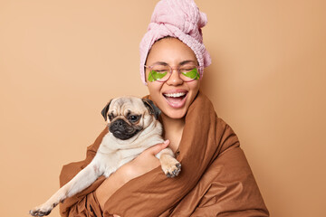 Positive dark skinned woman laughs happily wrapped in blanket poses with pug dog undergoes beauty...