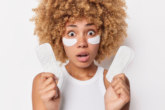 Shocked scared woman with blonde curly hair stares bugged eyes holds sanitary napkins applies beauty patches under eyes for skin treatment dressed in casual t shirt isolated over white wall.