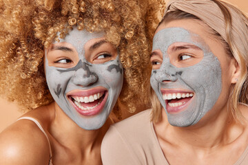 Close up shot of positive cheerful young women look at each other gladfully apply nourishing clay masks smile broadly undergo beauty procedures want to look beautiful on party. Skin care concept
