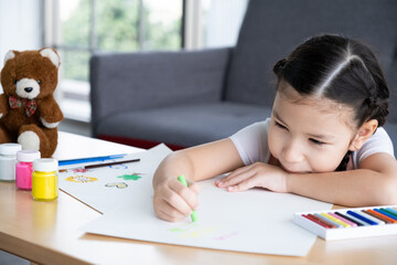 ํYoung girl sit on the sofa while using water color and crayon to paint on the paper inside of the living room. Children creativity and learning process, art and education for kids