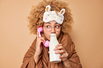 Pensive woman has conversation via stationary phone drinks fresh milk from bottle has breakfast wrapped in blanket wears sleepmask on forehead spectacles isolated over brown studio background