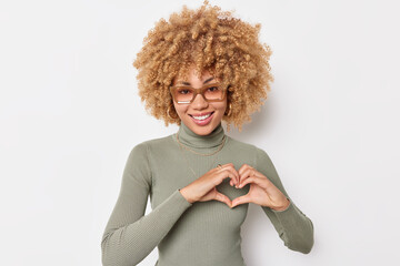 Pretty curly haired woman makes heart gesture over chest expresses love smiles gently wears...