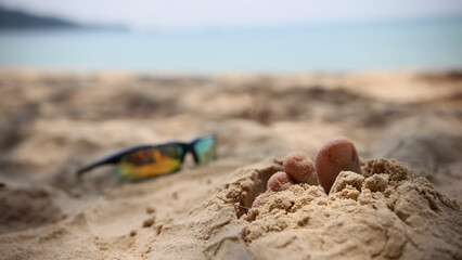 Tropical beautiful beach with sand, foot sticking out of the sand and sunny sunglasses. Summer travel or vacation concept.