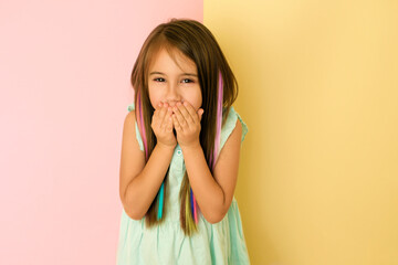 Long-haired girl with multicolored strands of curls cover  mouth with  hands laughing, trying to keep laughing. Kid on a pink-yellow background keeps secret. Artificial strands of colored hair in baby