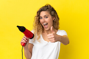 Young blonde woman holding a hairdryer isolated on yellow background with thumbs up because something good has happened