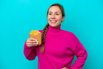 Young caucasian woman holding fried chips isolated on blue background posing with arms at hip and smiling