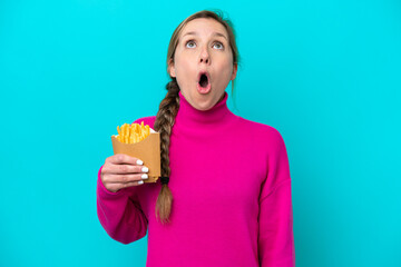 Young caucasian woman holding fried chips isolated on blue background looking up and with surprised expression