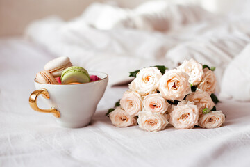 Sweet dessert, vanilla macaroons in mug and white roses on bed background. Romantic morning breakfast at bed.
