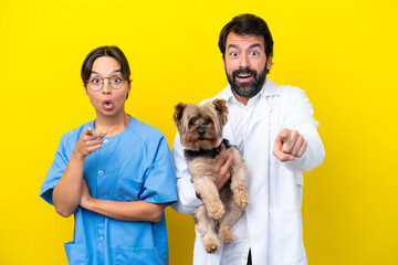 Young veterinarian couple with dog isolated on yellow background surprised and pointing front