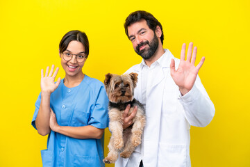 Young veterinarian couple with dog isolated on yellow background saluting with hand with happy expression