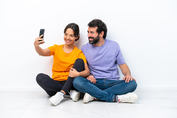 Fototapeta na wymiar Young caucasian couple sitting on the floor isolated on white background making a selfie with the mobile