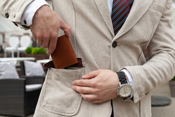 A stylishly dressed business man puts a brown leather wallet in his jacket pocket. Horizontal orientation, no face  - obrazy, fototapety, plakaty
