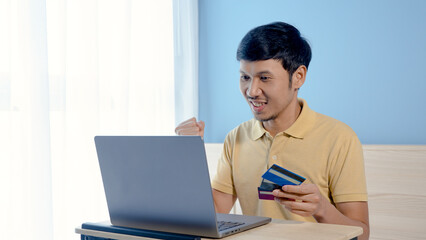 Handsome Asian man wearing a yellow shirt pays for online shopping on his laptop computer. The young man looked happy after completing his credit card payment.online shopping
