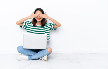 Young Vietnamese woman with a laptop sitting on the floor isolated on white wall covering eyes by hands