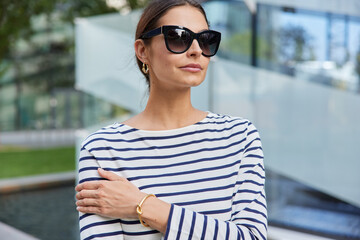 Horizontal shot of thoughtful European woman wears sunglasses casual striped jumper focused into distance walks outdoors poses at street against blurred background. Street lifestyle concept.