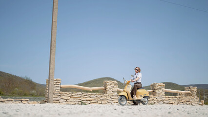 Young man on a yellow moped standing on a dusty countryside road on blue clear sky background....