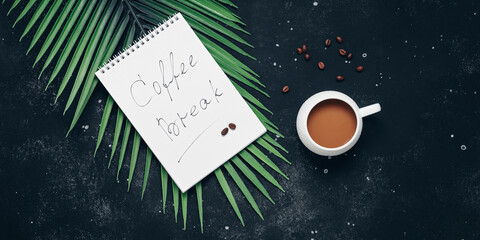 Coffee break. A cup of coffee with an open notepad and decorative palm leaves on a black stone background. Top view, banner.