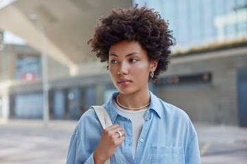 Outdoor shot of beautiful woman with dark curly hair wears blue casual shirt carries bag looks away strolls at street against blurred background during daytime. People and lifestyle concept.