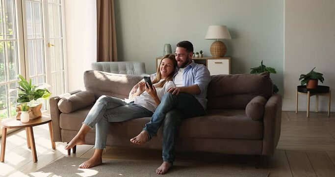 Relaxed couple sit on sofa use cellphone discuss purchase, make order, buying goods online, search house decorations ideas on internet spend carefree weekend with modern tech, fun and leisure concept