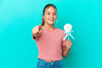 Little Caucasian girl holding a lollipop isolated on blue background surprised and pointing front