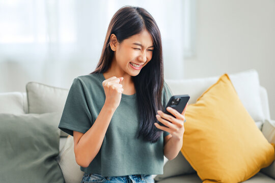 Excited young asian woman hold phone chatting in dating app feel happy sit on sofa at home, Closeup joyful female reading good news on phone. Surprised lady celebrating victory on phone