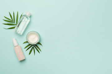 Facial tonic and cream with green leaves on color background, top view