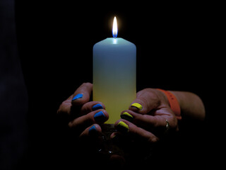 Płonąca świeca trzymana w kobiecych dłoniach. Świeca ma odcień niebieski i żółty. Na takie same kolory pomalowane są paznokcie. Jest to symbol solidarności z ofiarami wojny w Ukrainie,