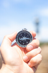 Closeup, hand holding an analog compass in front of a lighthouse on the coast, summer concept, holidays, travelling. Selective focus, vertical shot.