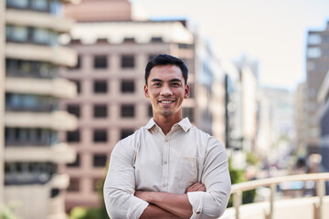 A young attractive Asian man stands in front of city buildings with arms crossed